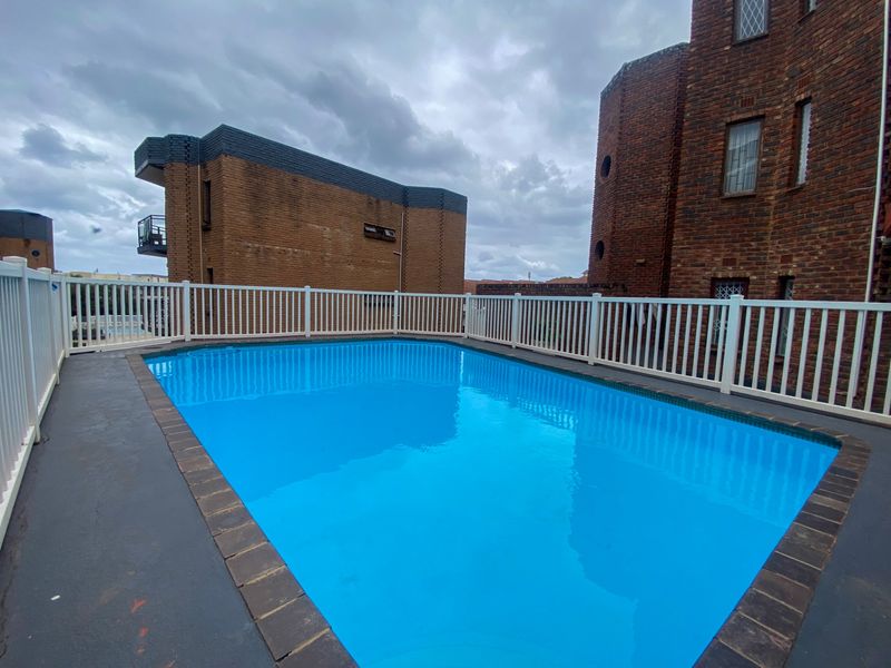 A communal swimming pool area featuring a clear blue pool surrounded by white safety fencing and brick apartment building exteriors.