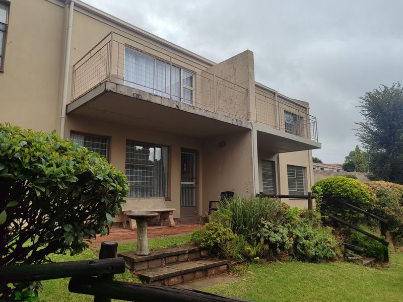 Rear view of the apartment building featuring a ground floor patio, upper level balcony with metal railings, and established garden area.