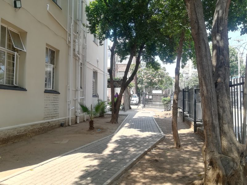 A paved walkway leads along the side of the apartment building, featuring security fencing and established trees in a shared outdoor space.