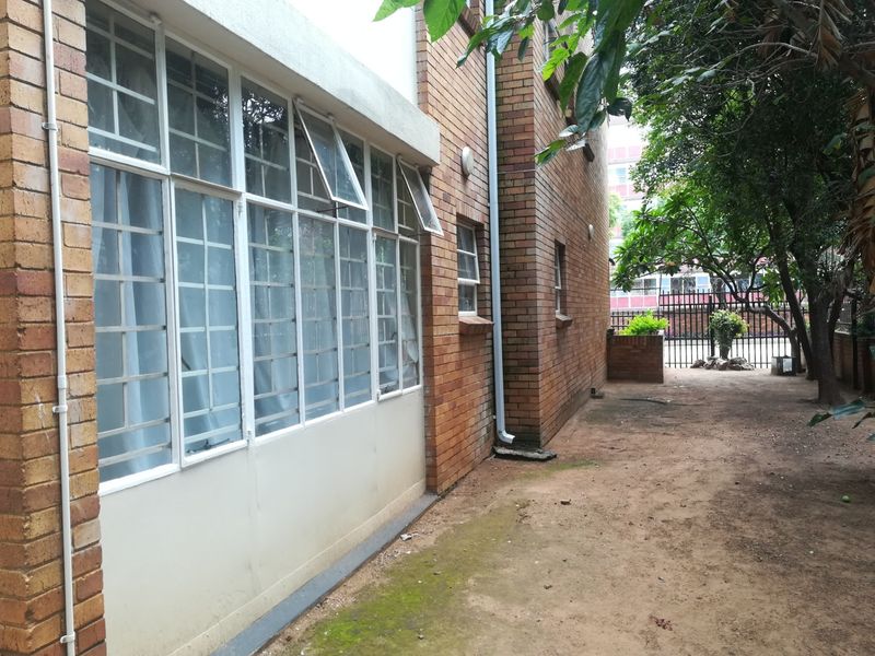 Rear exterior view of the apartment building featuring face brick walls, metal window frames with security bars, and an unpaved ground area.