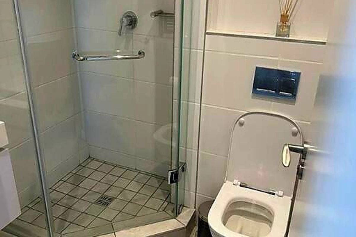 A compact modern bathroom featuring a glass-enclosed shower, wall-mounted toilet with chrome flush plate, and neutral floor-to-ceiling tiling.