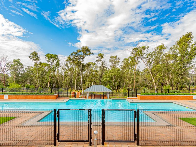 A well-maintained communal swimming pool area featuring paved decking, secure perimeter fencing, and a central gazebo set against a natural treed backdrop.