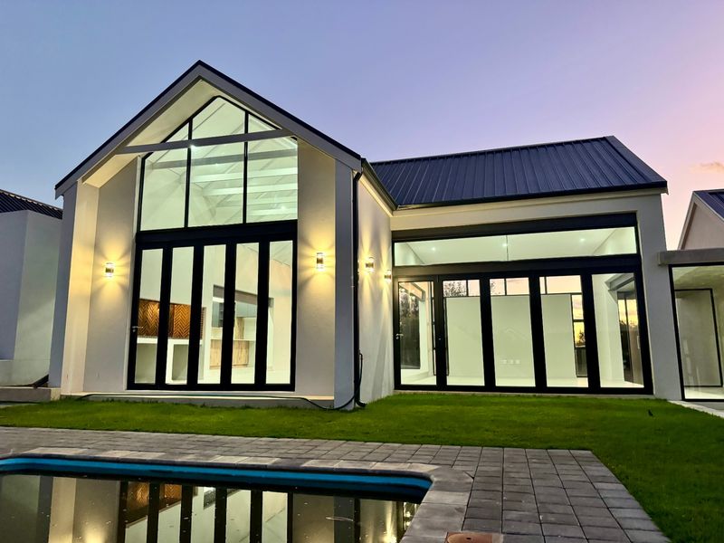 Modern rear elevation featuring a gabled roof, expansive glass doors, integrated wall lighting, and a paved patio area overlooking a swimming pool.