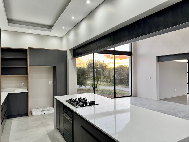 Modern open-plan kitchen featuring sleek dark cabinetry, stone countertops, integrated gas hob and oven, and expansive windows with scenic sunset views.