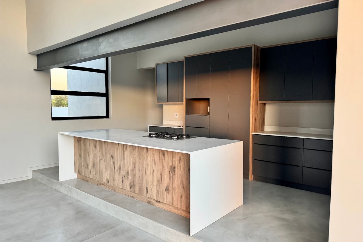 Modern kitchen featuring a central island with a gas hob, sleek dark cabinetry, stone countertops, and polished concrete floors under an exposed steel beam.