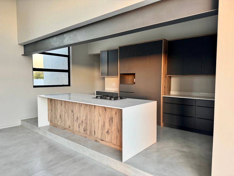 Modern kitchen featuring a central island with a gas hob, sleek dark cabinetry, stone countertops, and polished concrete floors under an exposed steel beam.