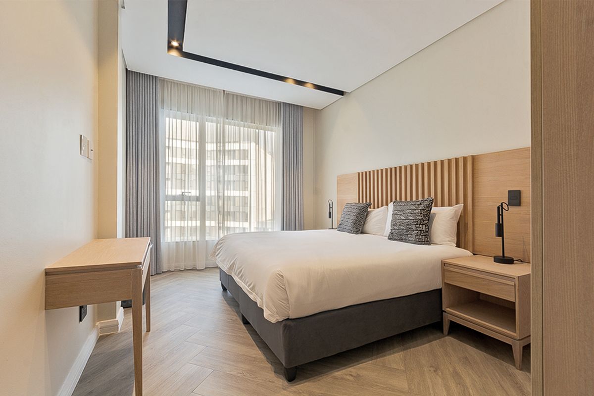 Modern bedroom featuring herringbone wood-look flooring, a custom slatted timber headboard, recessed ceiling lighting, and floor-to-ceiling window treatments.
