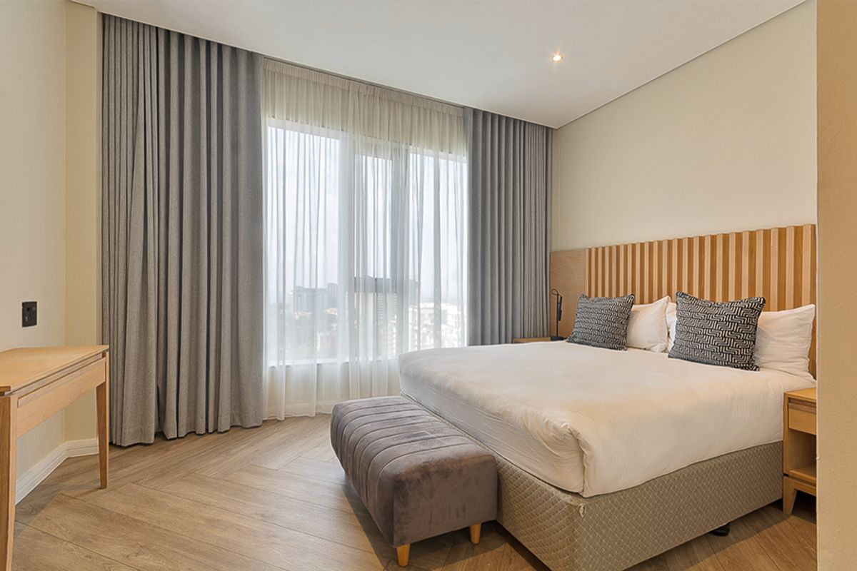 A modern, well-appointed bedroom featuring herringbone timber flooring, a stylish timber slat headboard, and floor-to-ceiling curtains with natural light.
