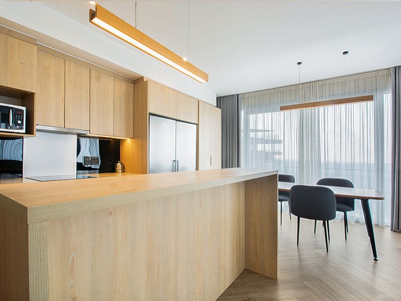 Modern open-plan kitchen and dining area featuring light timber cabinetry, integrated appliances, a central island, and contemporary pendant lighting with floor-to-ceiling window access.