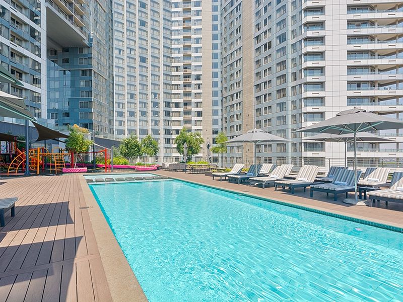 A well-maintained outdoor swimming pool area featuring timber decking, sun loungers, and an adjacent children's playground set against a high-rise apartment complex.