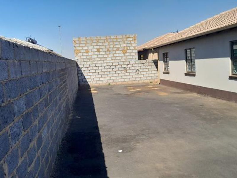 Rear exterior view showing a paved yard area, concrete block boundary wall, and the side elevation of the house with tiled roof.