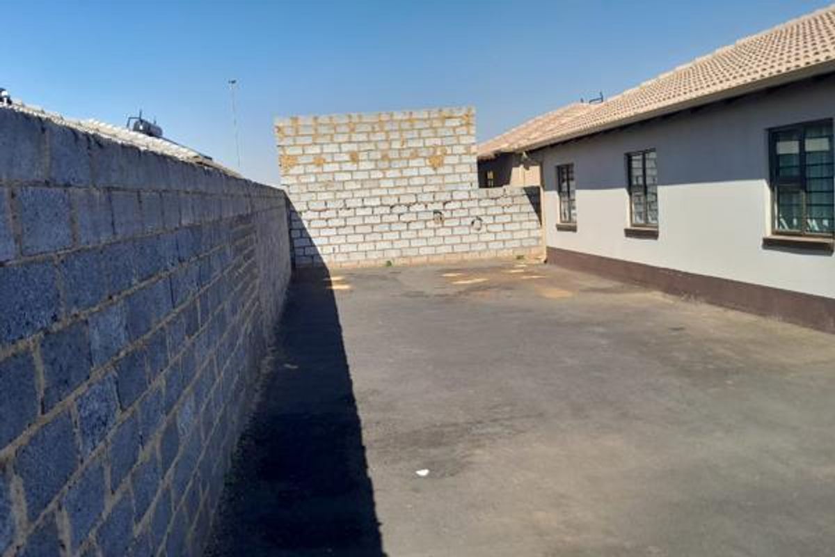 Rear exterior view showing a paved yard area, concrete block boundary wall, and the side elevation of the house with tiled roof.