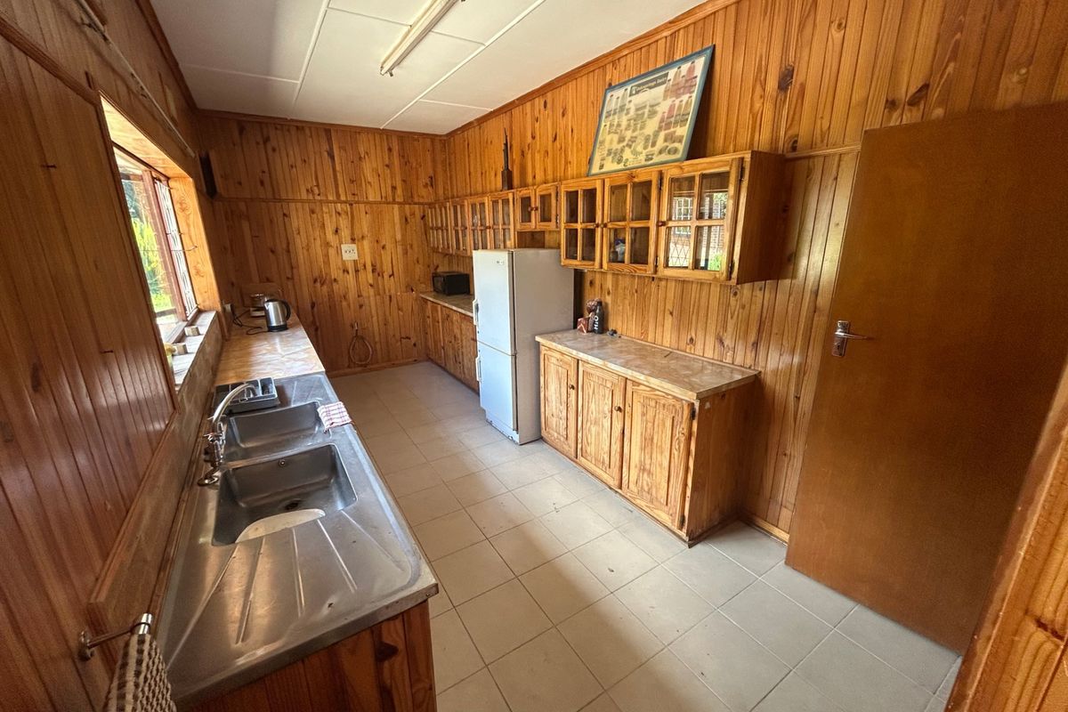 A dated kitchen featuring extensive timber wall paneling, wooden cabinetry, laminate countertops, and a double bowl stainless steel sink with tiled flooring.
