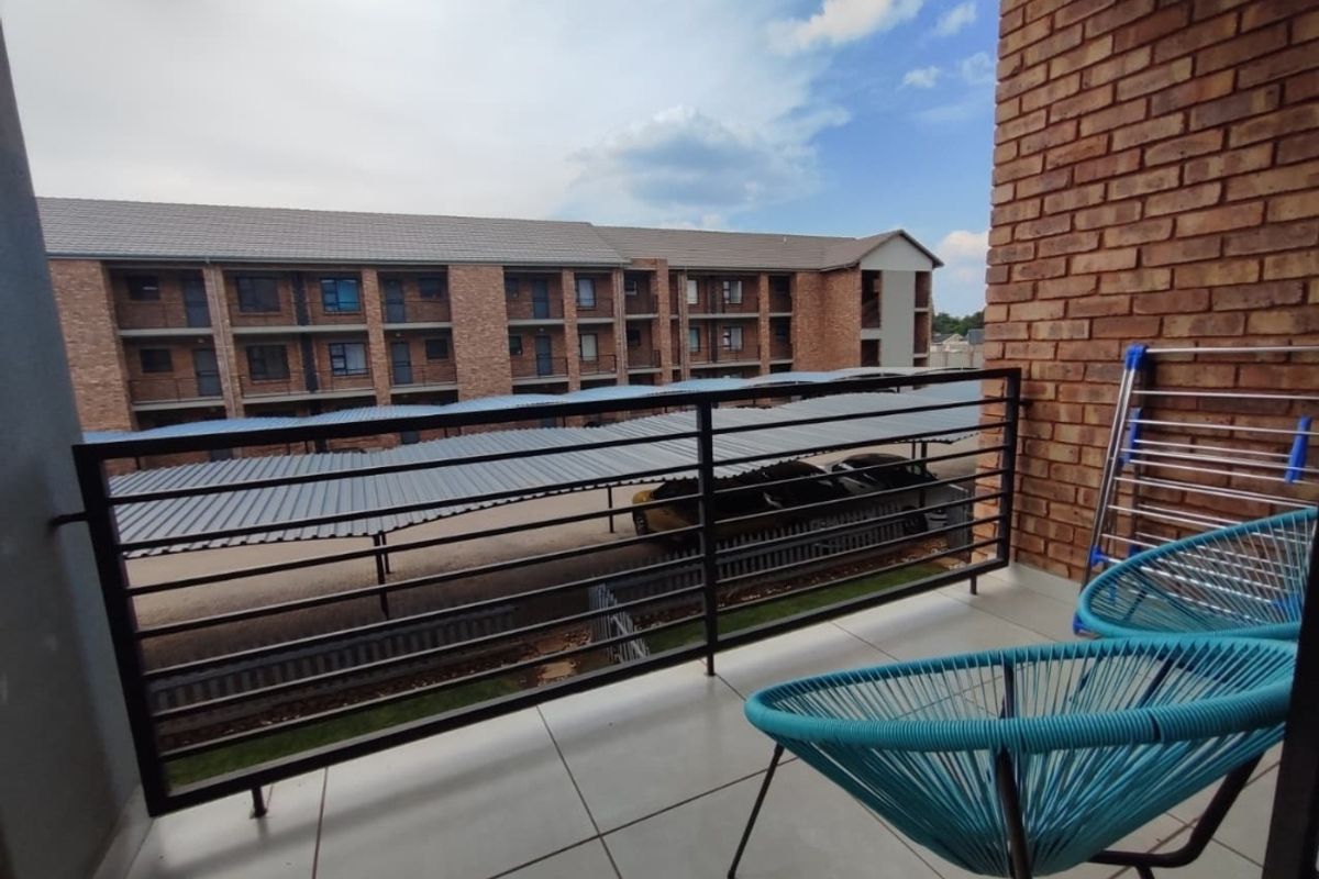 A private balcony featuring tiled flooring, a metal balustrade, and an exposed brick wall, overlooking the apartment complex parking area.