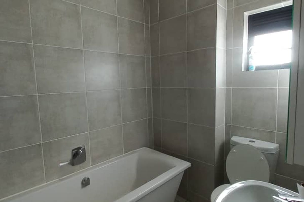 A modern bathroom featuring neutral grey wall tiles, a built-in bathtub, a standard toilet, and a washbasin with natural light from a window.