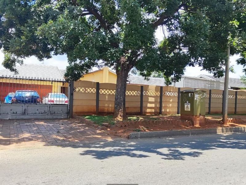 Street view of the property featuring a paved driveway, secure metal gate, and a precast concrete boundary wall with mature trees.