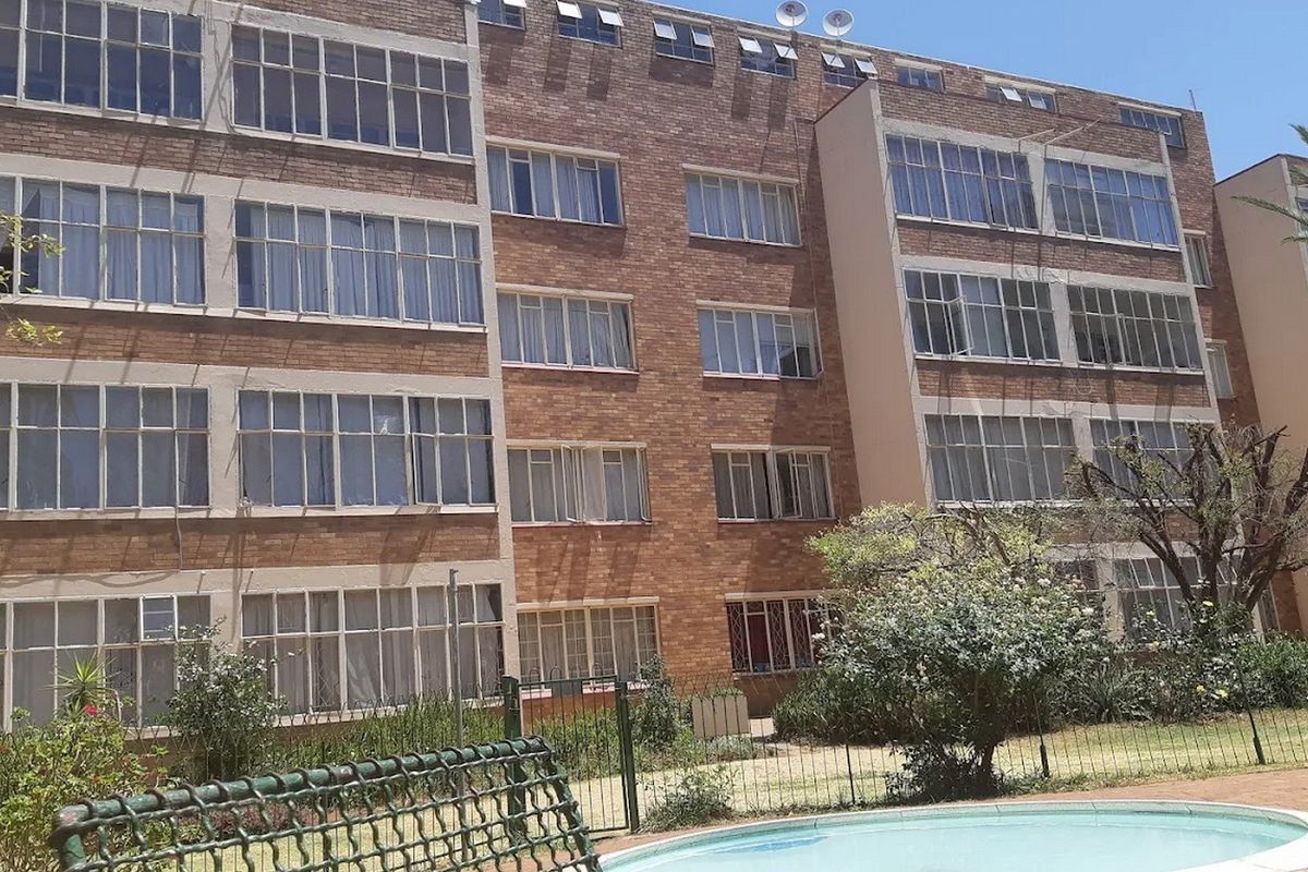 Exterior view of a multi-story brick apartment building featuring large steel-framed windows, overlooking a communal swimming pool and garden area.