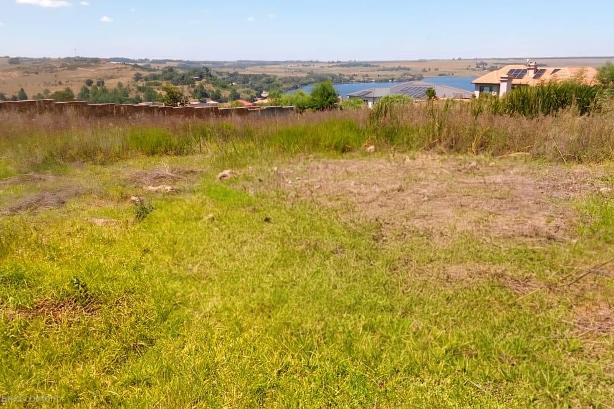 A vacant residential plot featuring natural grass terrain, perimeter boundary wall, and scenic views of the surrounding landscape and nearby water body.