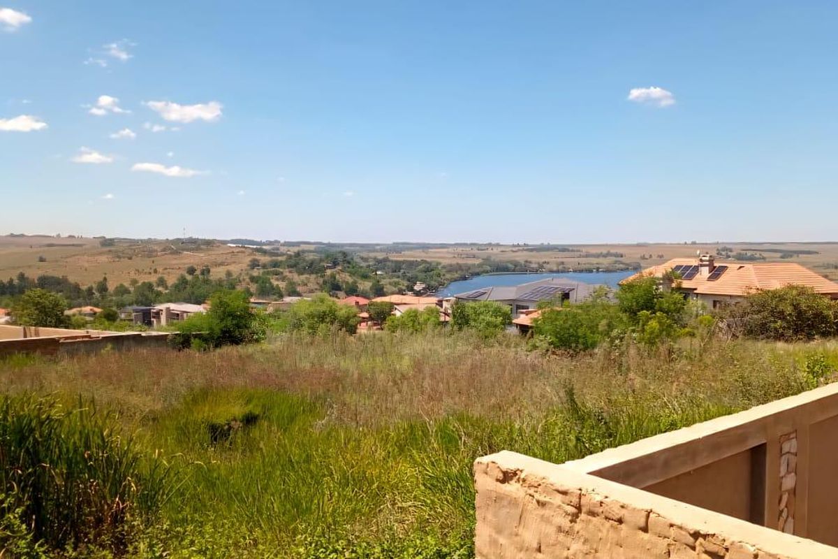 A vacant residential plot featuring expansive landscape views, including a nearby lake and surrounding estate housing, with an existing partial boundary wall.