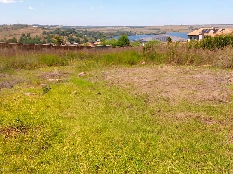 A vacant residential plot featuring natural grass terrain, perimeter boundary wall, and scenic views of the surrounding landscape and nearby water body.