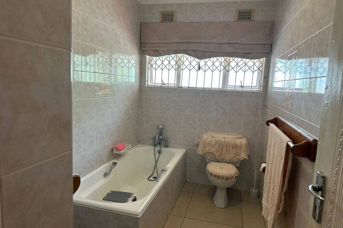 A functional bathroom featuring a built-in bathtub, toilet, and full-height ceramic wall tiling with security bars on the window.