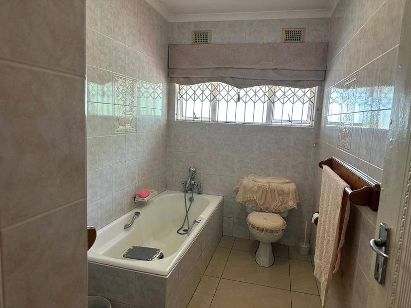 A functional bathroom featuring a built-in bathtub, toilet, and full-height ceramic wall tiling with security bars on the window.