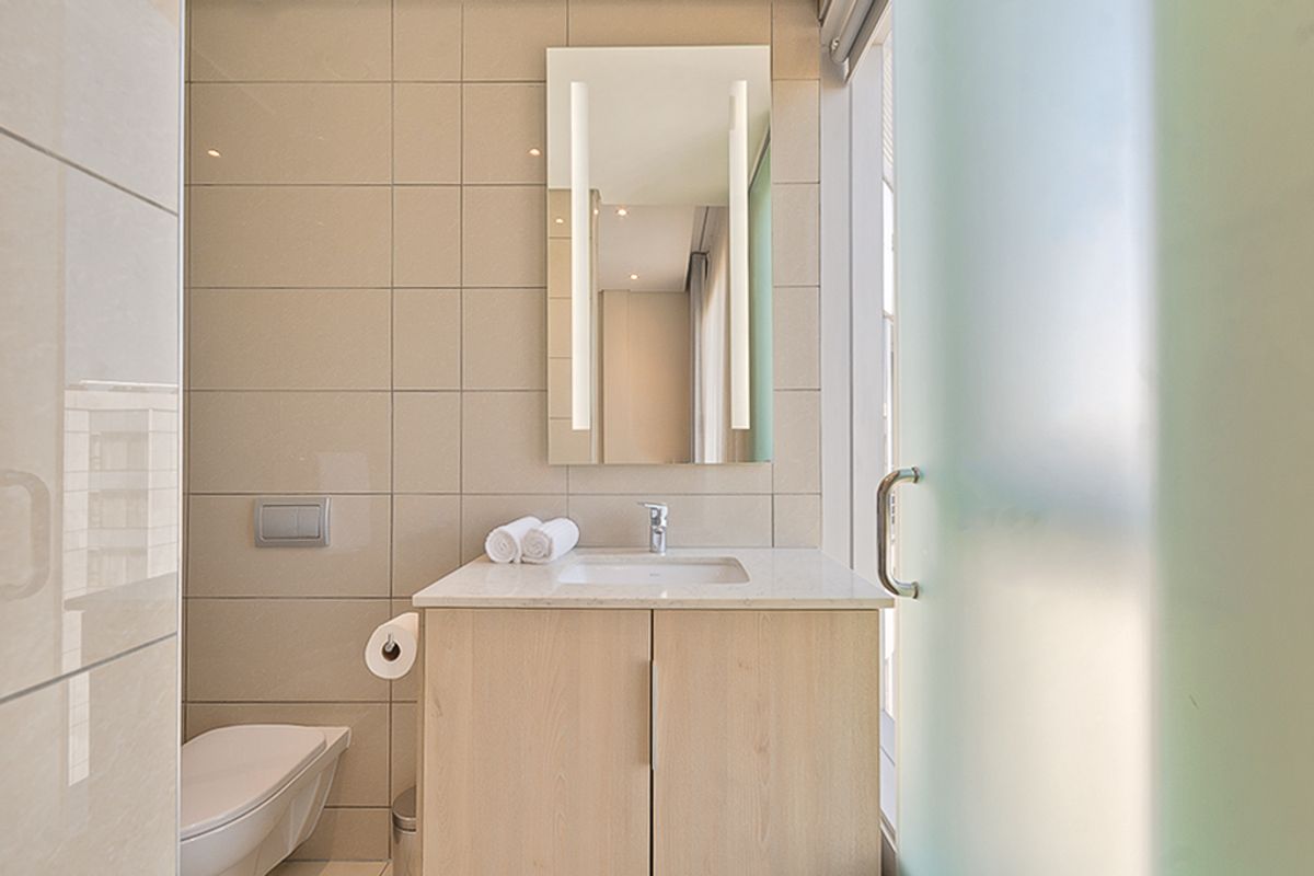 Modern bathroom featuring a wall-mounted toilet, light wood vanity with stone countertop, integrated mirror lighting, and a frosted glass door.