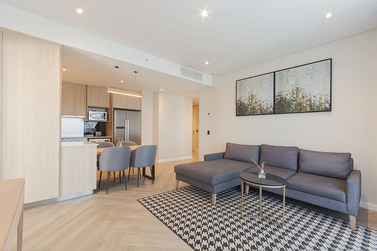 Modern open-plan living area featuring light wood cabinetry, herringbone flooring, recessed lighting, and integrated kitchen appliances in a well-maintained apartment setting.