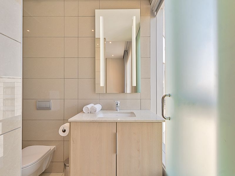 Modern bathroom featuring a wall-mounted toilet, light wood vanity with stone countertop, integrated mirror lighting, and a frosted glass door.