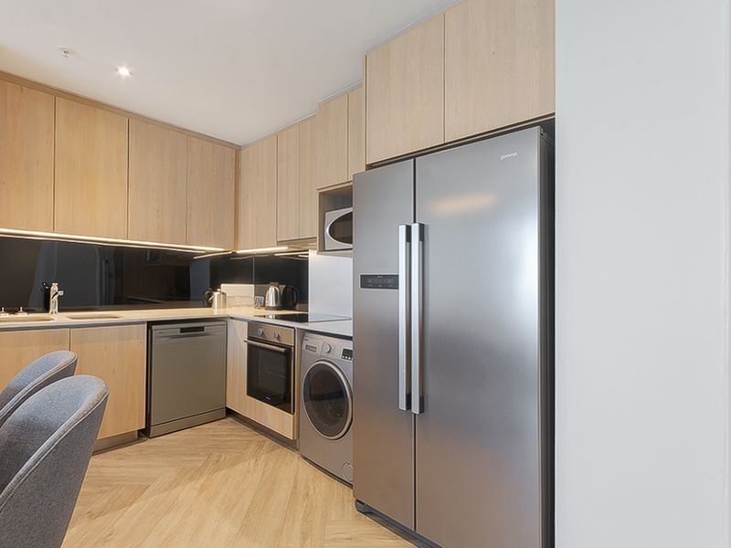 Modern kitchen featuring light wood cabinetry, integrated appliances, sleek dark splashback, and herringbone flooring, offering a clean and contemporary aesthetic.