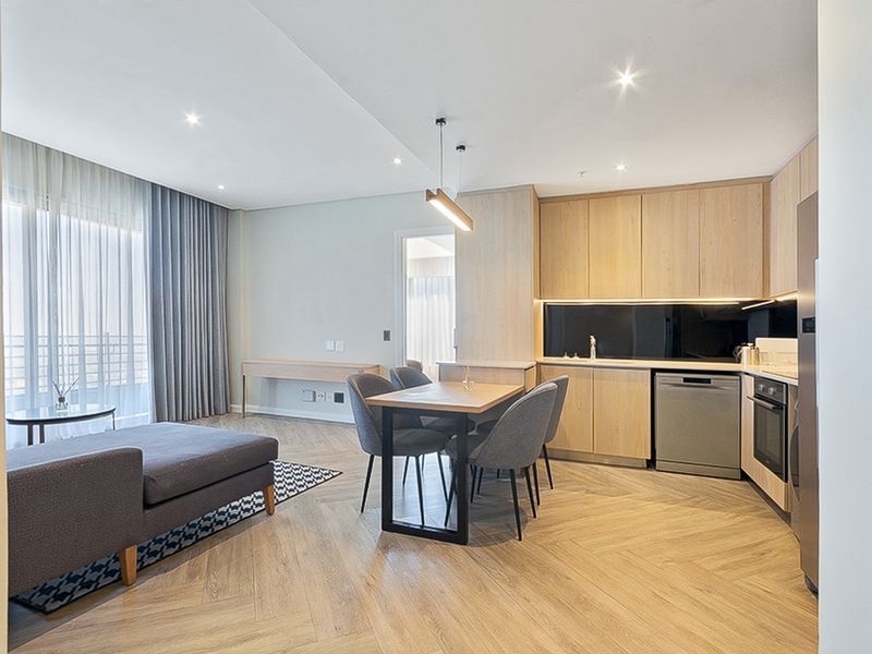 Modern open-plan living area featuring herringbone wood flooring, sleek light wood cabinetry, integrated kitchen appliances, and direct access to a balcony through sliding doors.