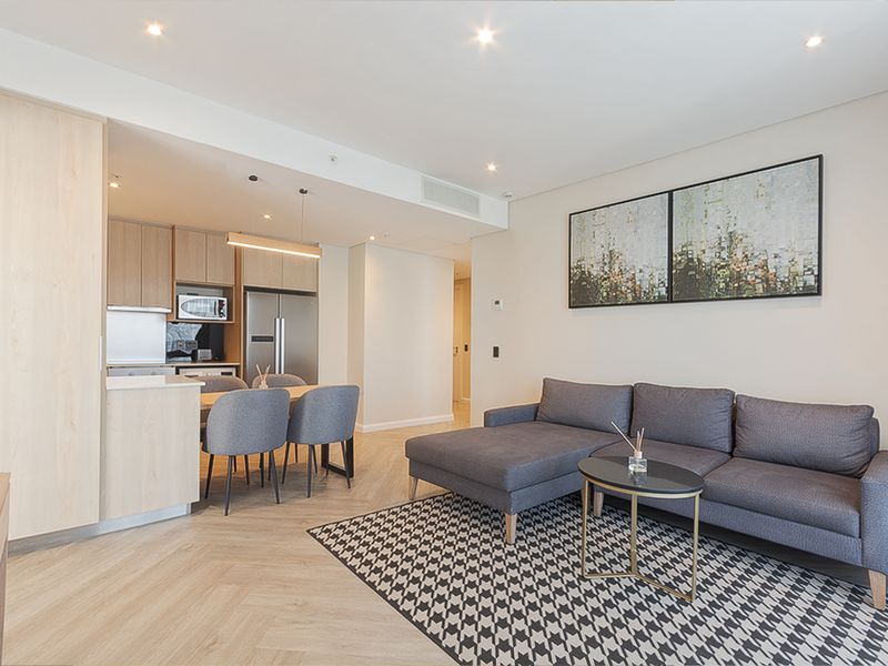 Modern open-plan living area featuring light wood cabinetry, herringbone flooring, recessed lighting, and integrated kitchen appliances in a well-maintained apartment setting.