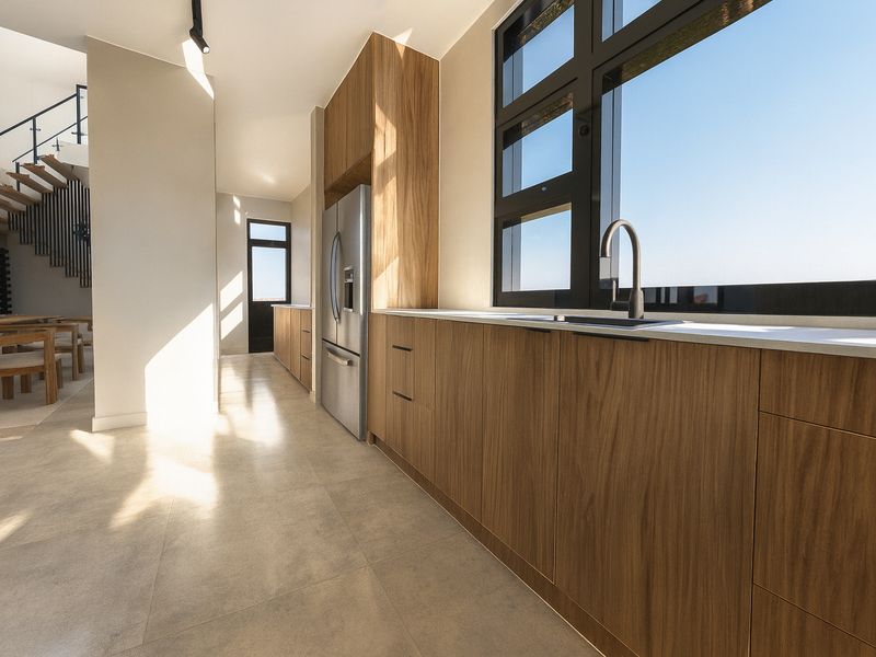 Modern open-plan kitchen featuring wood-grain cabinetry, stone countertops, and large windows, seamlessly flowing into the dining area with a feature staircase.