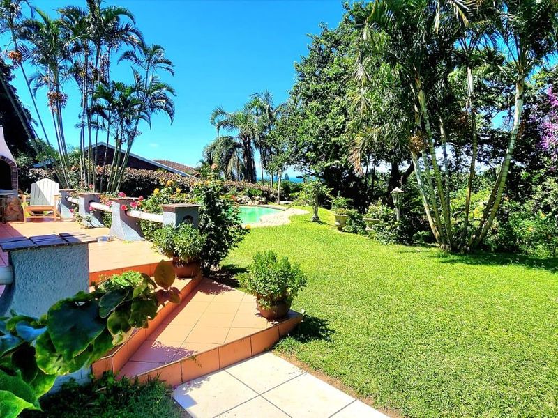 A spacious tiled patio featuring a built-in braai and retractable awning, overlooking a lush garden and swimming pool area.