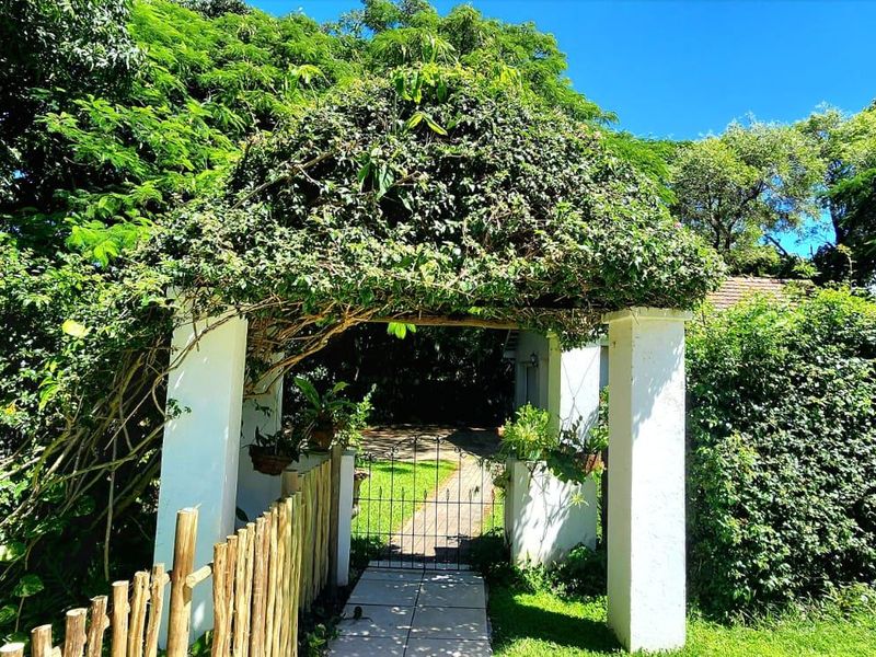 A charming garden entrance featuring a masonry archway with climbing greenery, a wrought iron gate, and a paved pathway leading into the property.