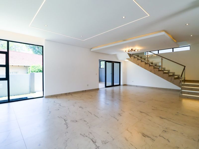 Spacious open-plan living area featuring modern marble-look floor tiles, recessed ceiling lighting, and a contemporary staircase with glass balustrades and integrated step lighting.