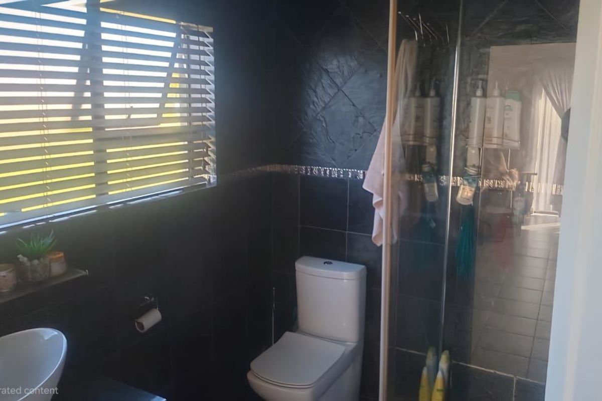 A bathroom featuring dark wall tiles, a white toilet, a glass-enclosed shower, and a window with horizontal blinds.