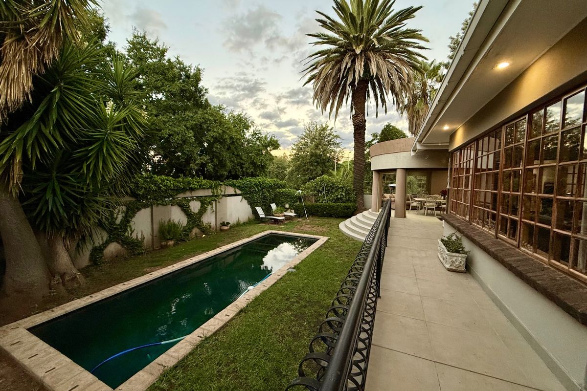 A well-maintained rear garden featuring a rectangular swimming pool, paved patio area, and established landscaping with a large palm tree.