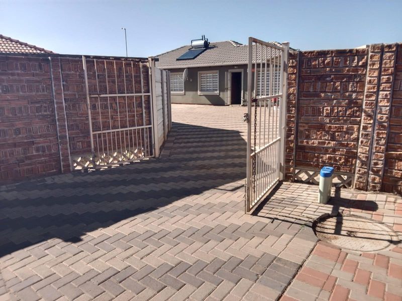 Front entrance view of a residential property featuring a paved driveway, secure metal gate, and brick boundary walls with a solar water heater installed.