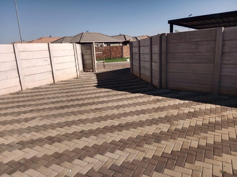 A paved driveway leading to a gated entrance, enclosed by precast concrete boundary walls with an adjacent carport structure.