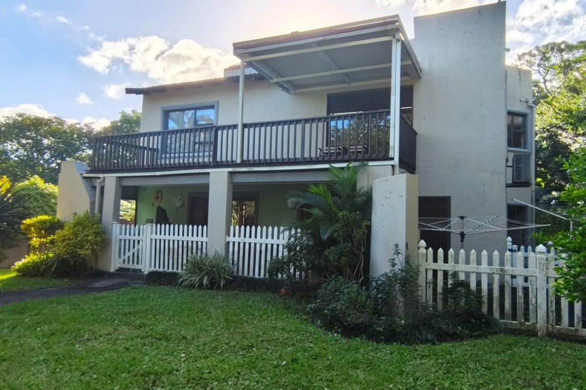 Rear view of a two-story house featuring a covered balcony, ground-level patio, and a well-maintained garden with white picket fencing.