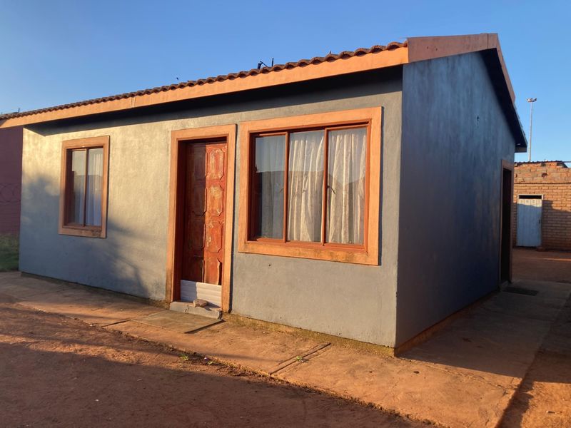 A modest single-story residential dwelling featuring a tiled roof, timber entrance door, and sliding windows with a surrounding concrete apron.