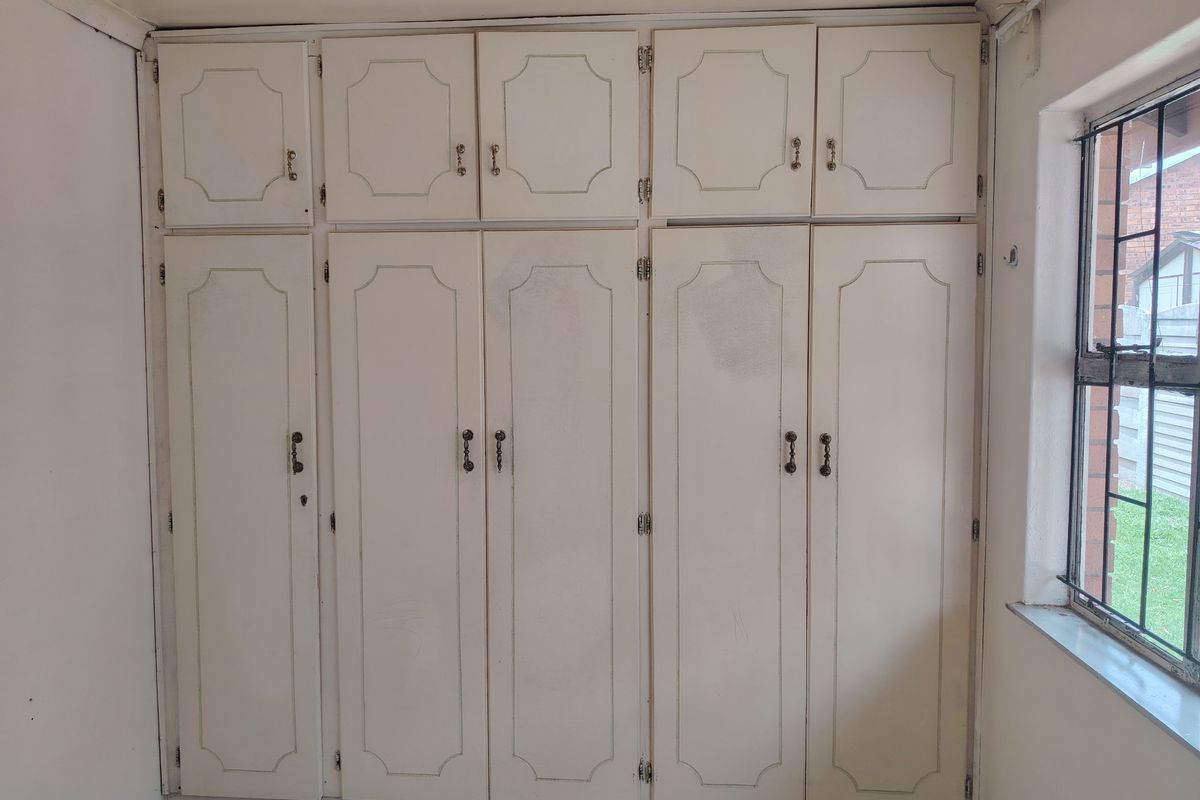 A bedroom featuring a large wall of dated built-in cupboards and a steel-framed window, offering a blank canvas for renovation.