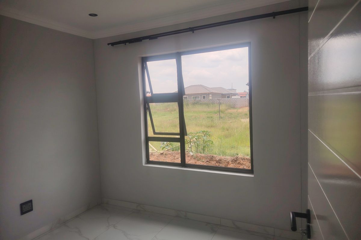 A bright, empty bedroom featuring modern white marble-look floor tiles, neutral grey walls, and a large aluminium-framed window with a view of the surrounding area.