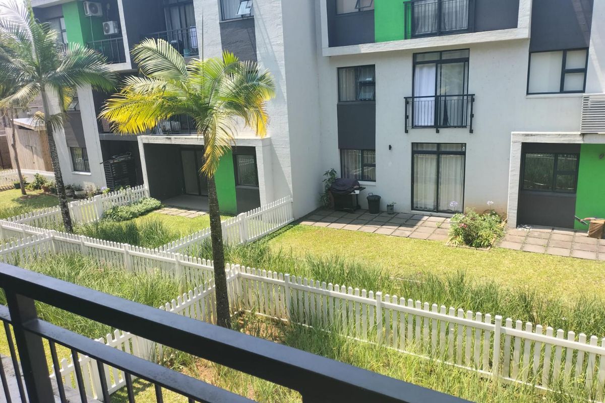 Rear view of the apartment complex featuring ground floor paved patios, private garden areas enclosed by white picket fencing, and upper-level balconies.