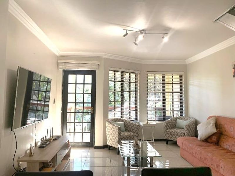 A bright living room featuring tiled flooring, coved cornicing, and a glass-paneled door leading to an outdoor area, complemented by large windows with venetian blinds.
