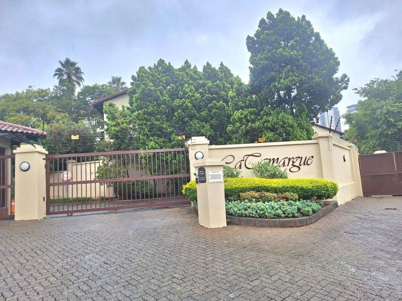 The secure entrance to La Camargue apartment complex, featuring a paved driveway, automated gate, and perimeter electric fencing.