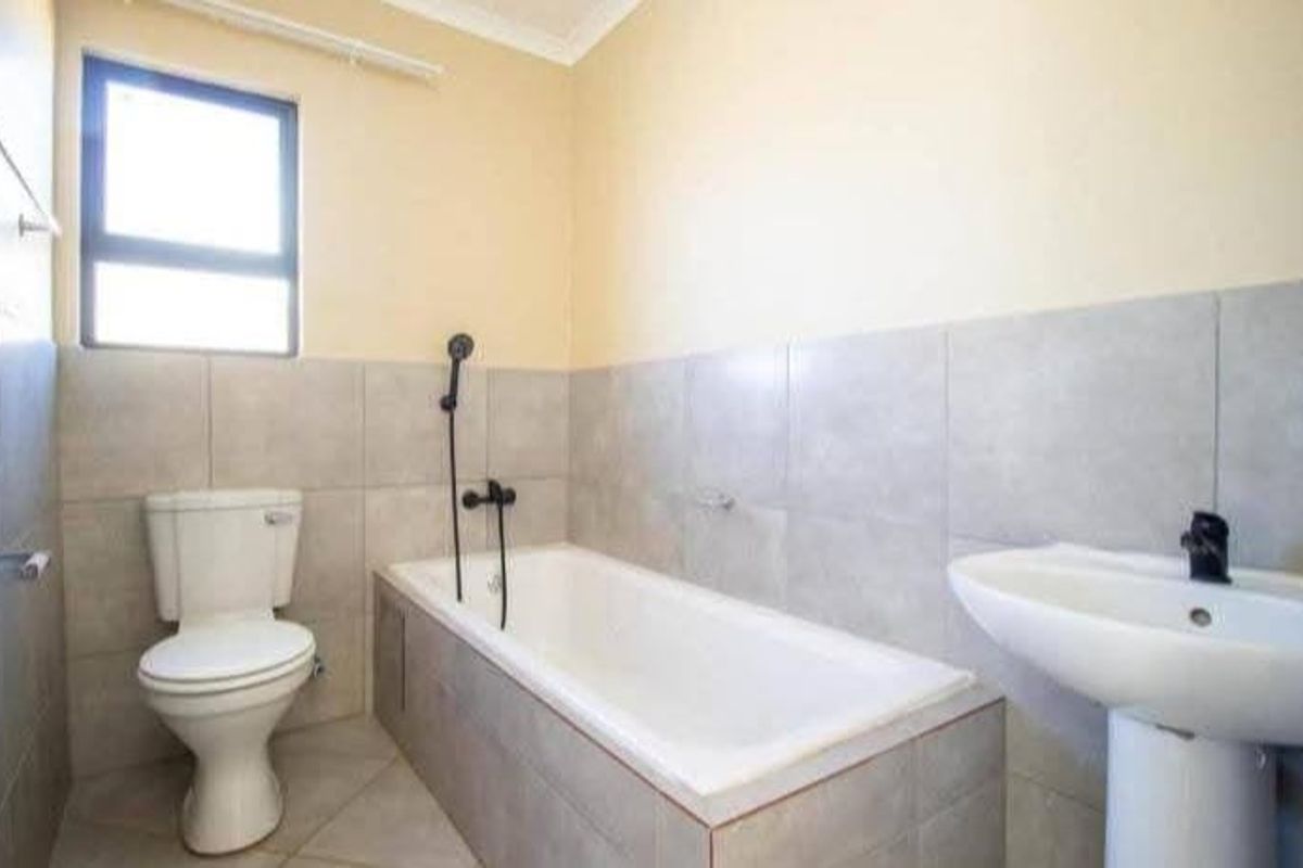 A functional bathroom featuring a bathtub with shower mixer, pedestal basin, and toilet, finished with neutral wall and floor tiling.