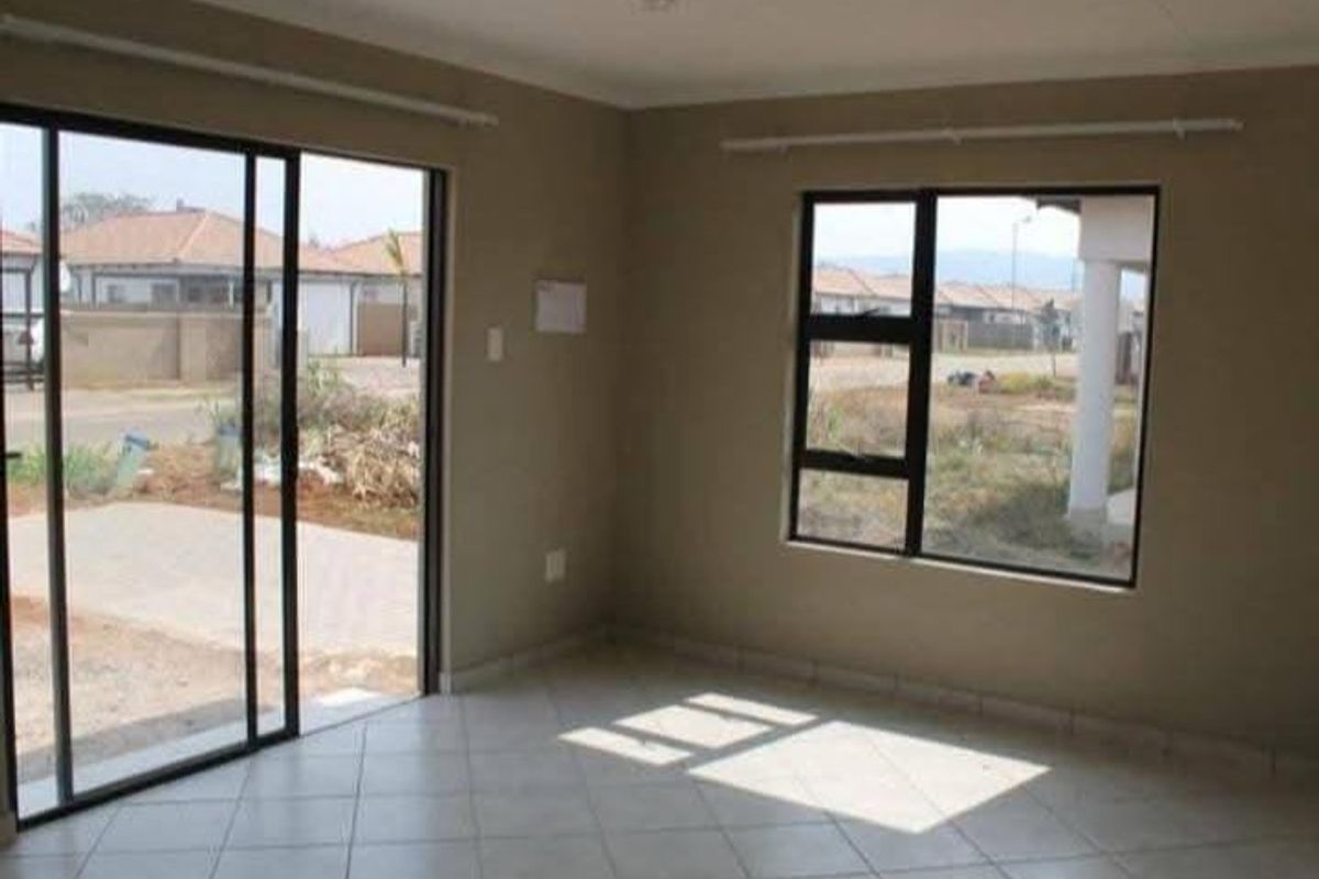 An empty, neutral-toned living room featuring tiled flooring, large aluminium-framed windows, and a sliding glass door providing access to the exterior.