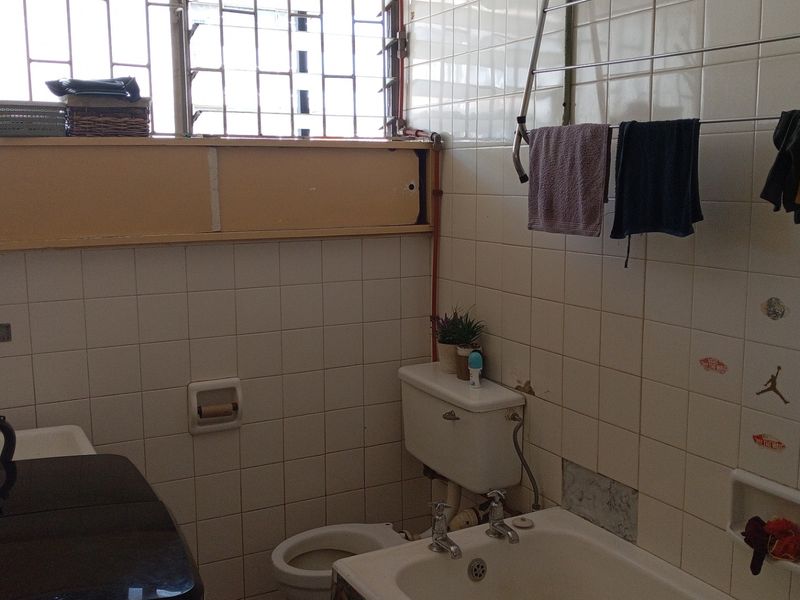 A dated bathroom featuring white ceramic wall tiles, a built-in bathtub, and a window with security grilles. The space requires cosmetic updates and modernization.